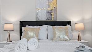 Una cama bien arreglada con almohadas blancas y doradas, dos lámparas de noche y un cuadro abstracto grande en la pared.