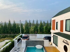 A modern building with a pool, two cars parked outside, and a view of trees and mountains in the background. - Verona Pool Villa (Pak Chong)
