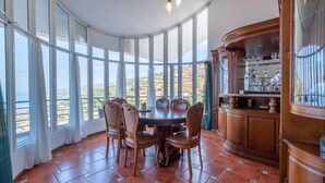 A dining area with a round table and chairs, large windows with curtains, and a wooden cabinet with glassware. - Casa Almada (Ribeira Brava)
