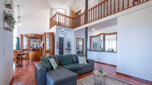 A spacious living area with a grey sectional sofa, a herringbone-patterned floor, a wooden staircase, and a kitchen visible in the background. - Casa Almada (Ribeira Brava)