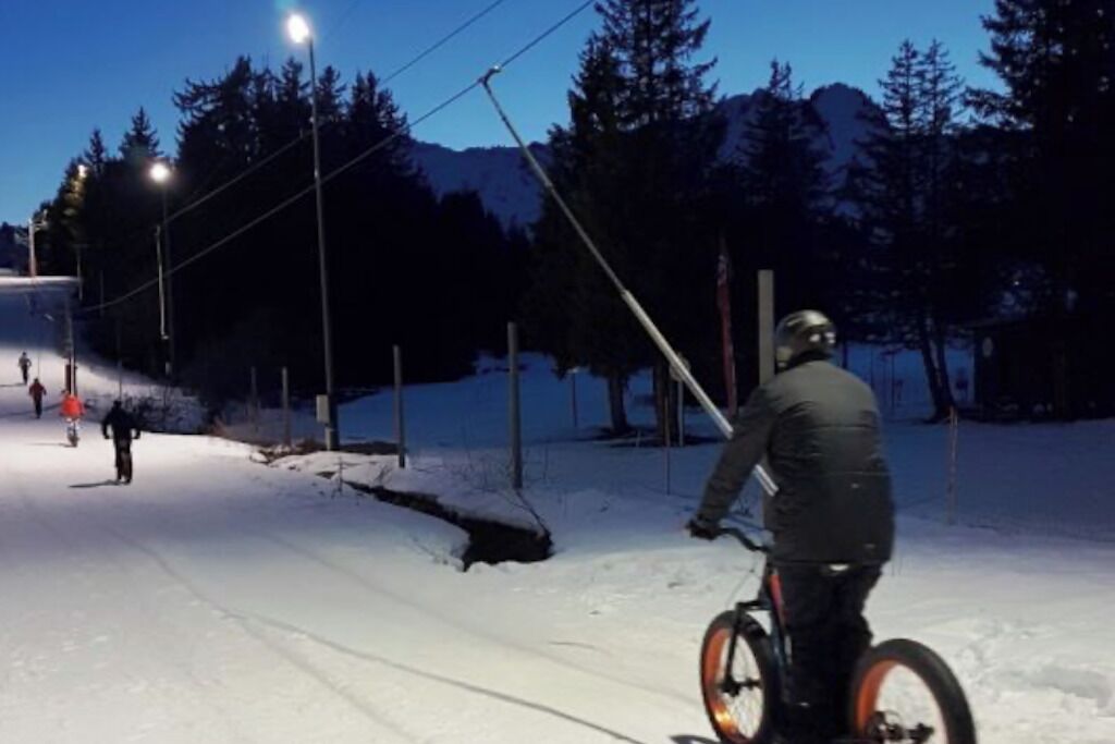 Eine Person fährt auf einem mit Schnee bedeckten Weg Fahrrad, im Hintergrund Straßenlaternen und Bäume.
