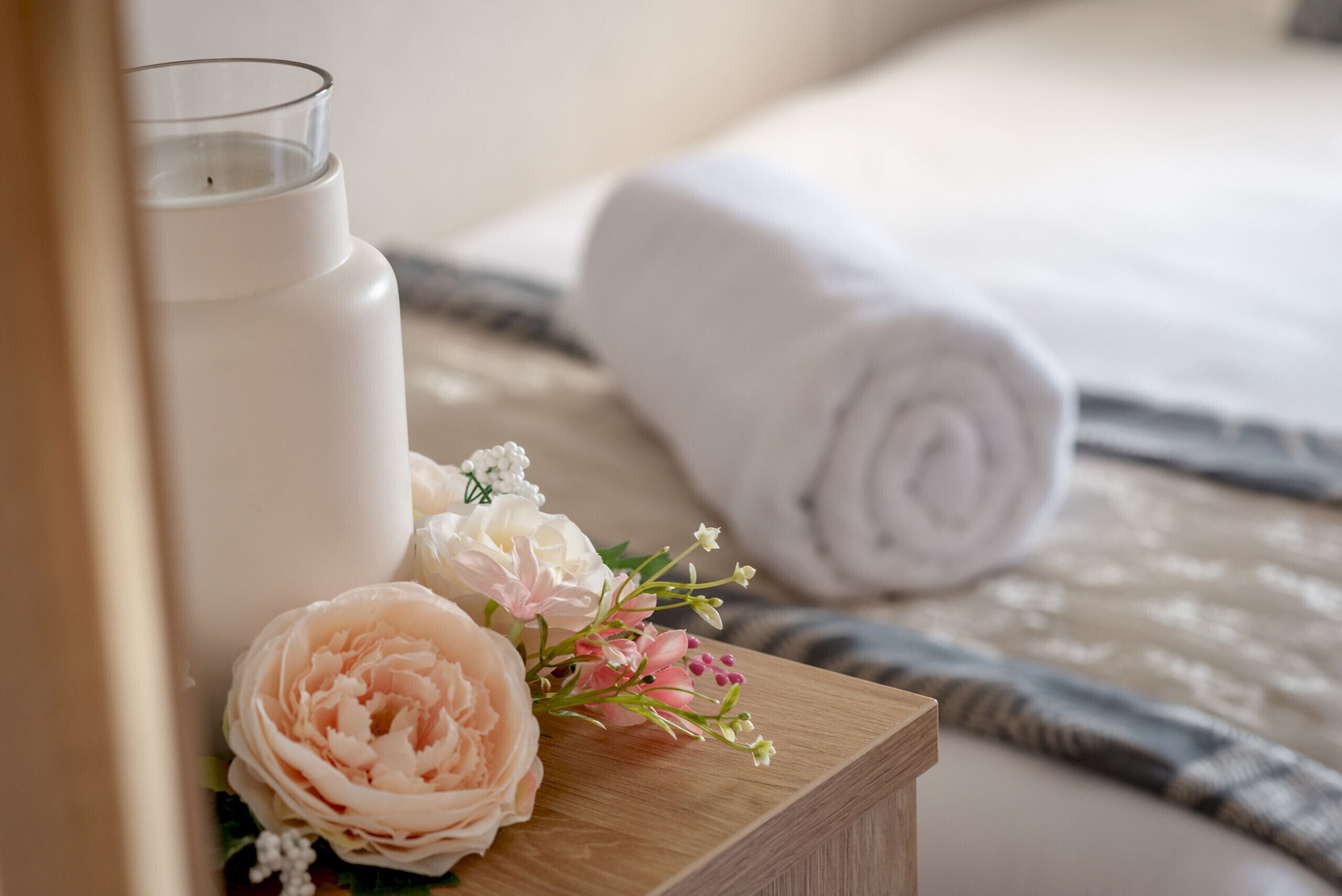 Une table de chevet en bois, sur laquelle se trouvent un porte-bougie en verre, un bouquet de fleurs et une serviette enroulée, le tout placé sur un lit recouvert d’une couvre-lit à motifs.