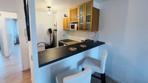 Une cuisine de taille réduite avec des meubles en bois, un plan de travail noir et un four à micro-ondes. On y trouve deux chaises blanches et une bouteille d’eau posée sur le plan de travail.