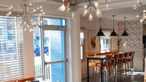 A modern dining area with multiple tables and chairs, a large window with blinds, and a ceiling with hanging light fixtures. - Stunning waterfront on bluff with sauna, firepit! (Weems)