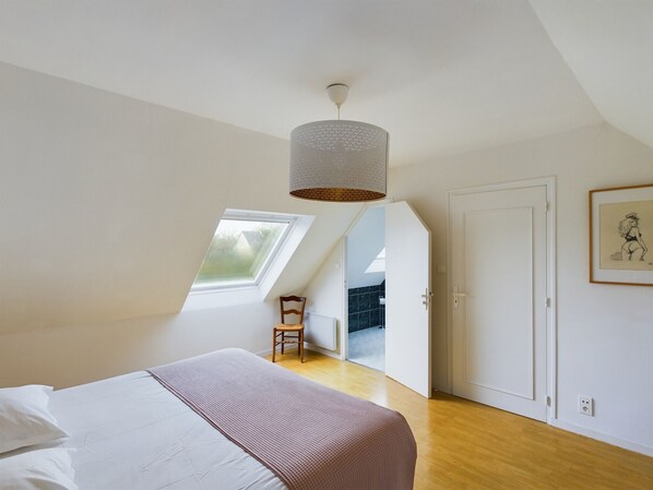 A bedroom with a bed, a chair, a ceiling light, a door, and a window. - -NAYA- Maison familiale à deux pas de la mer (Trégunc)