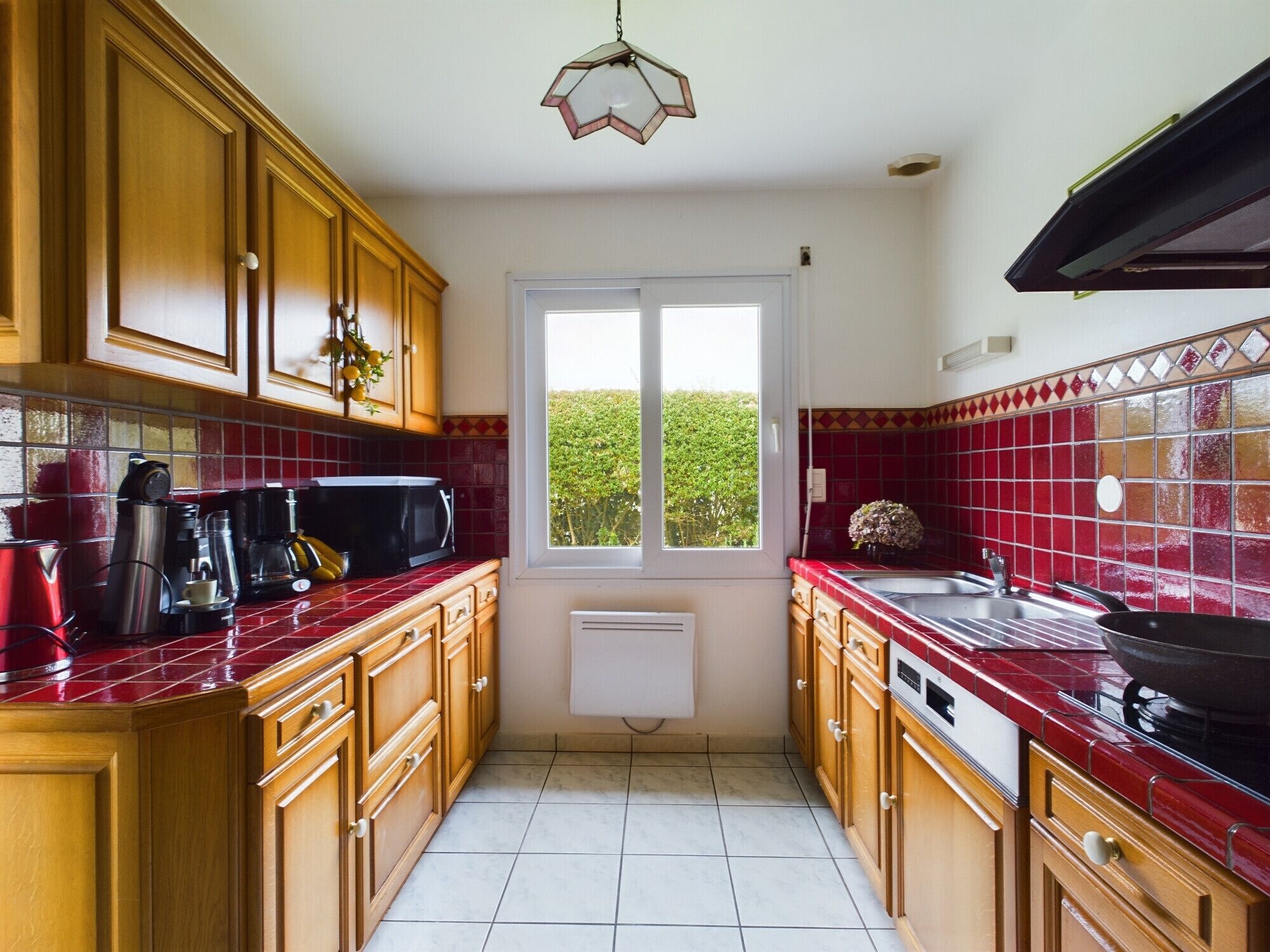 A compact kitchen with wooden cabinets, a red tiled backsplash, a window with a view, and a hanging light fixture.