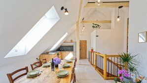 A dining area with a wooden table set for four, a kitchen in the background, and a staircase with wooden railings.