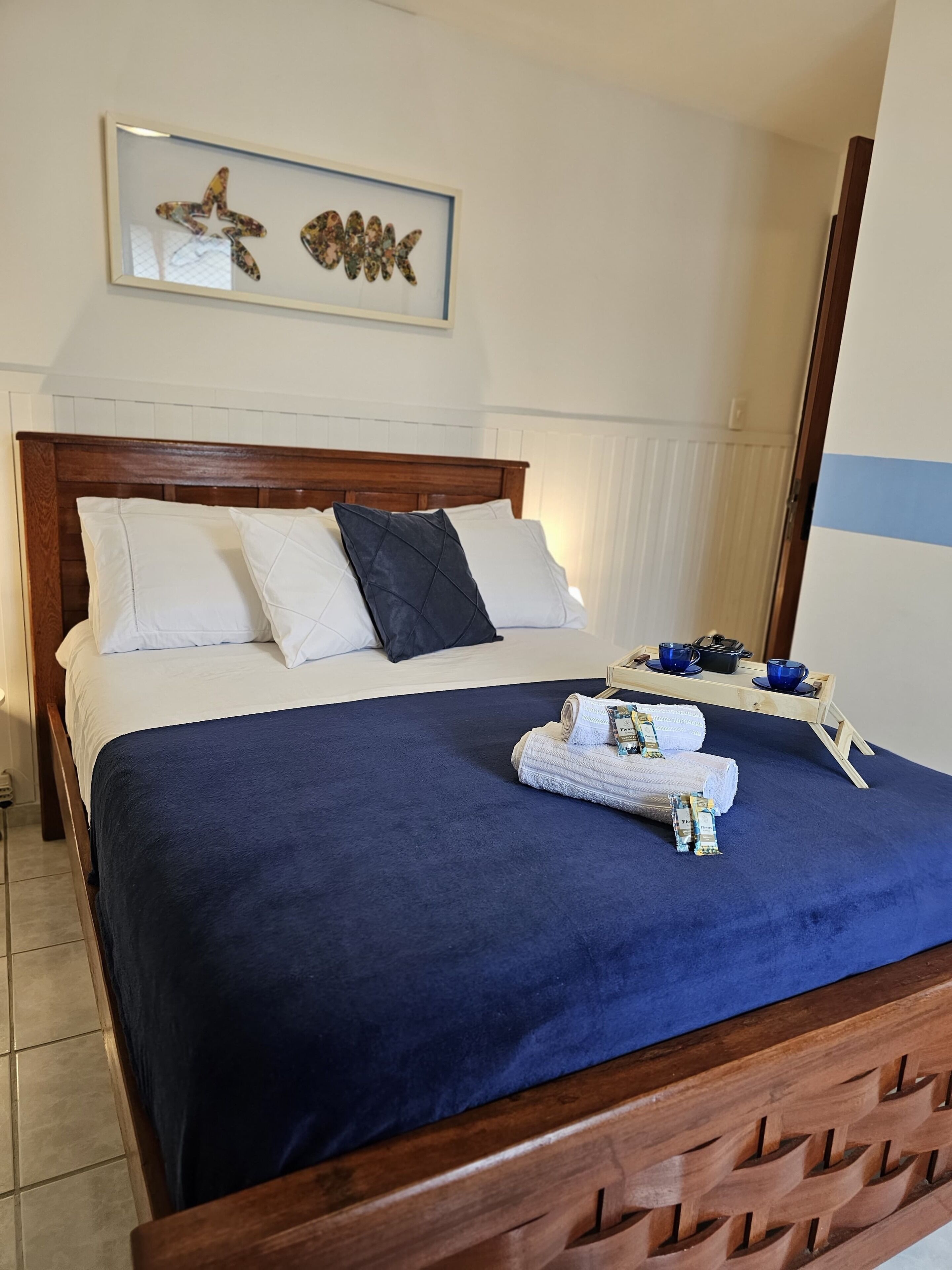 Un lit bien fait, recouvert d’une couvre-lit bleue, avec des oreillers blancs et un plaid bleu foncé. On y voit une tête de lit en bois, un tableau encadré au-dessus du lit, et un plateau en bois avec des tasses et une bouilloire sur le côté.