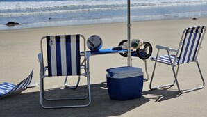 A beach scene with a blue and white striped beach umbrella, two striped beach chairs, a cooler, and a soccer ball on a metal table. - Comfort & Beautiful View 300m from Paradise - Wi-Fi/AC (Parnamirim)