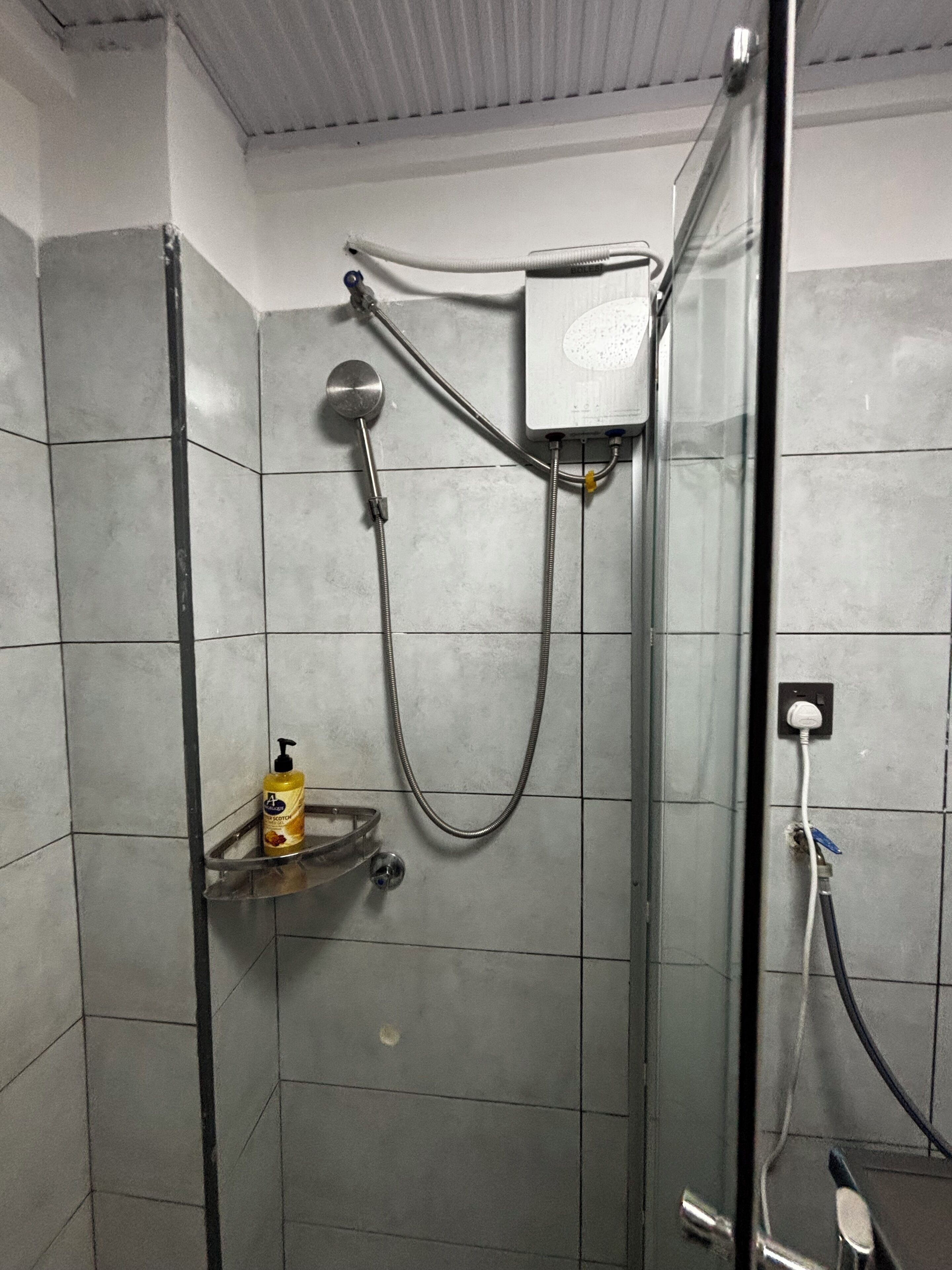 Dusche mit Handbrause, fest installierter Bedienung und Seifenspender auf einer kleinen Ablage.