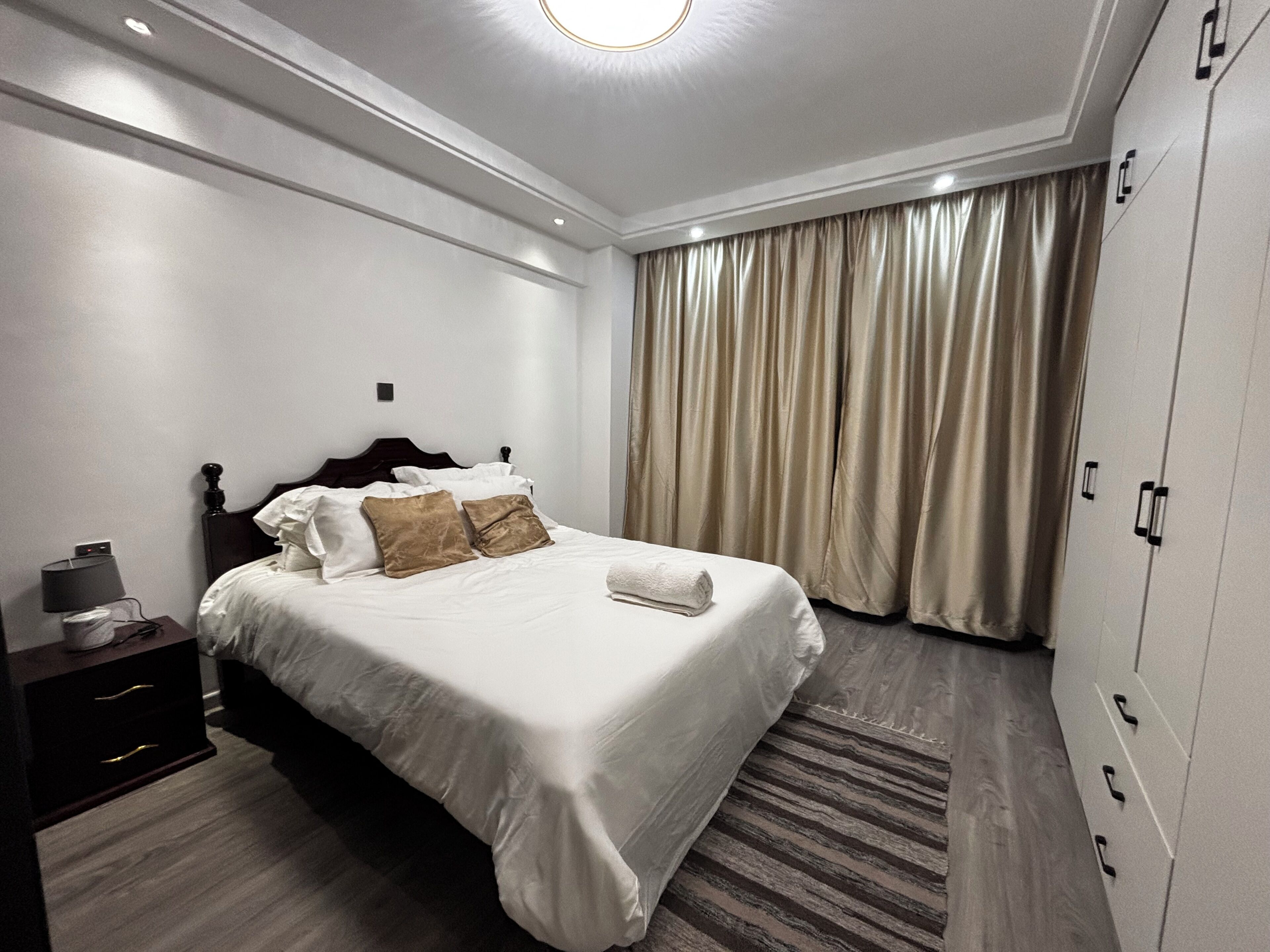 A neatly made bed with pillows, a bedside lamp, a wardrobe, and a curtain.