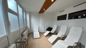 Une salle de spa avec quatre chaises longues blanches, un radiateur, une table avec des magazines et de grandes fenêtres.