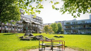 Une aire de jeux avec une balançoire circulaire, entourée d’une zone herbeuse parsemée de gros rochers et d’arbres. Au fond, on aperçoit des immeubles à plusieurs étages, dotés de balcons et d’un espace de repos abrité.