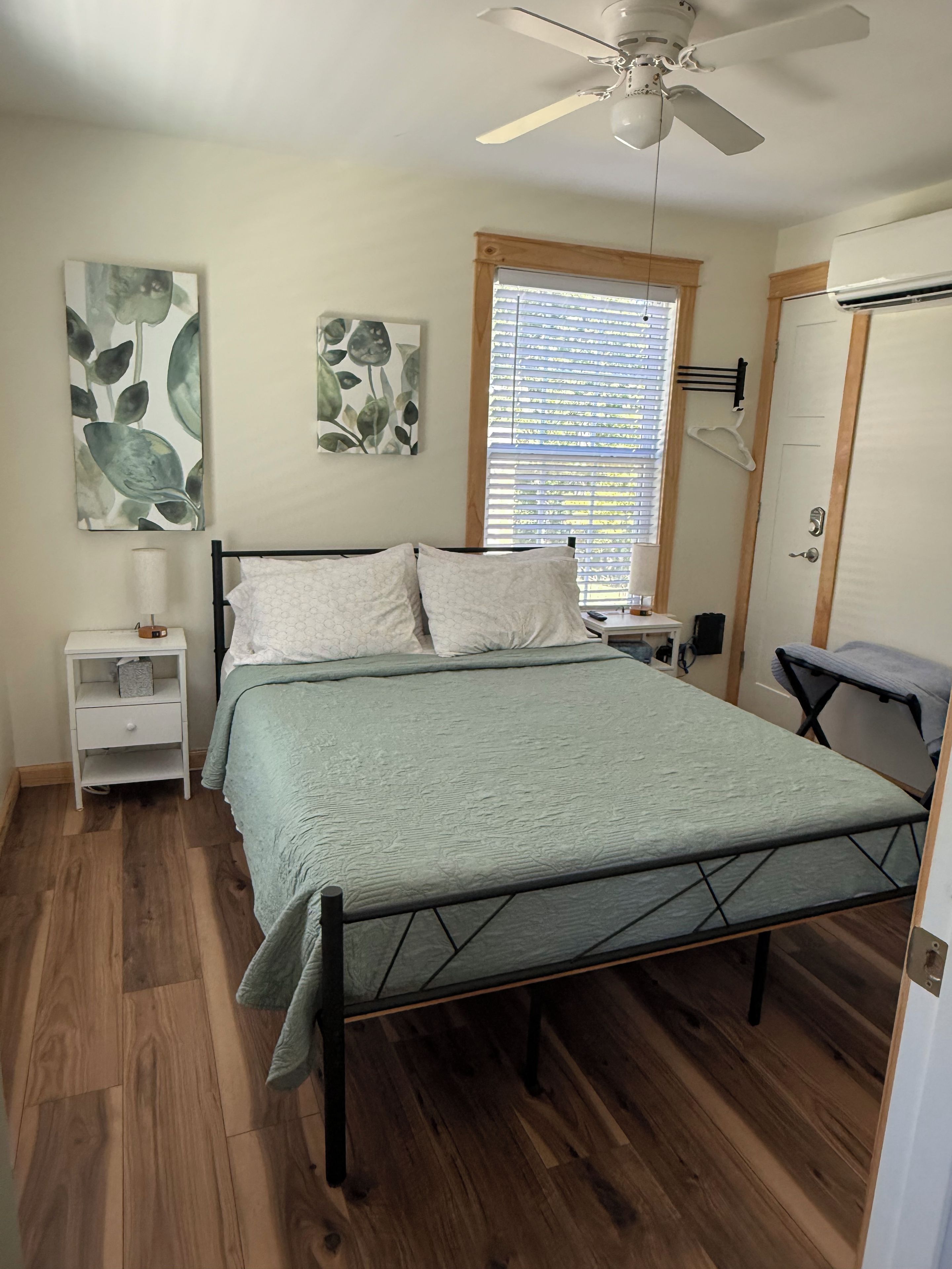 A bedroom with a bed, a nightstand, a ceiling fan, a window with blinds, and a door.
