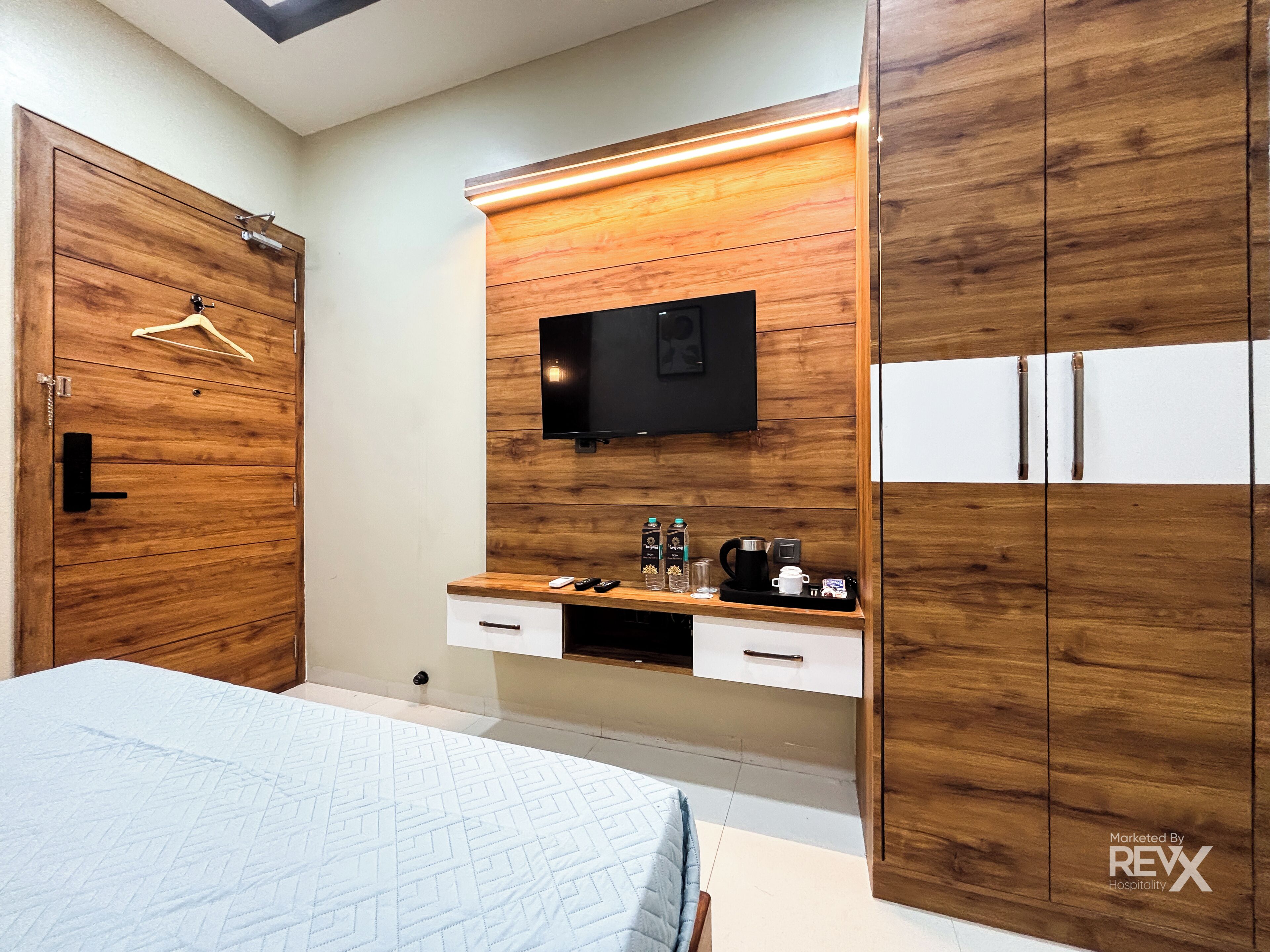 A bedroom with a bed, wooden wardrobe, a TV mounted on the wall, a small shelf with toiletries, and a bedside table.