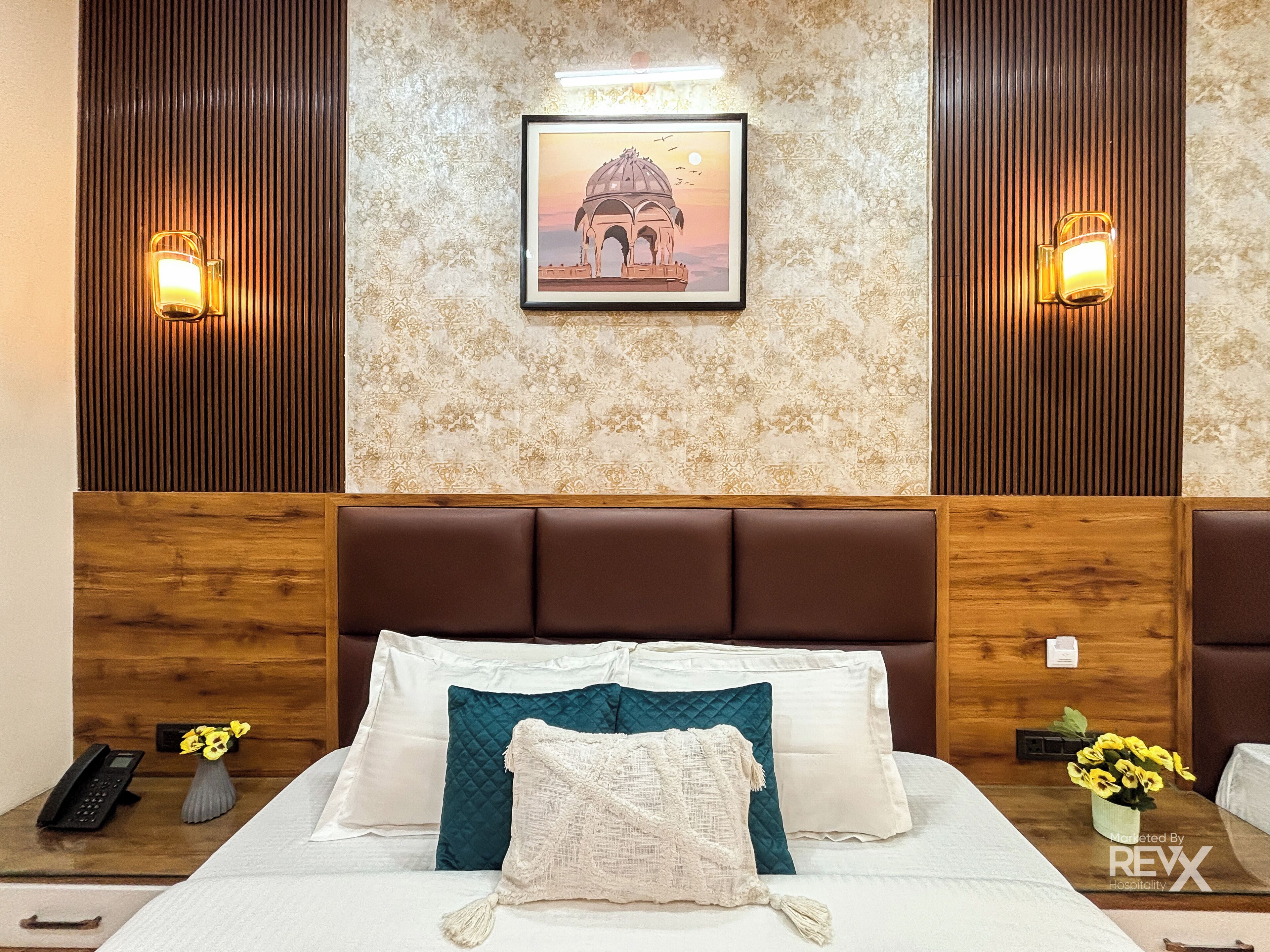 A hotel room with a large bed, a wooden headboard, a framed picture on the wall, and two wall-mounted lights.