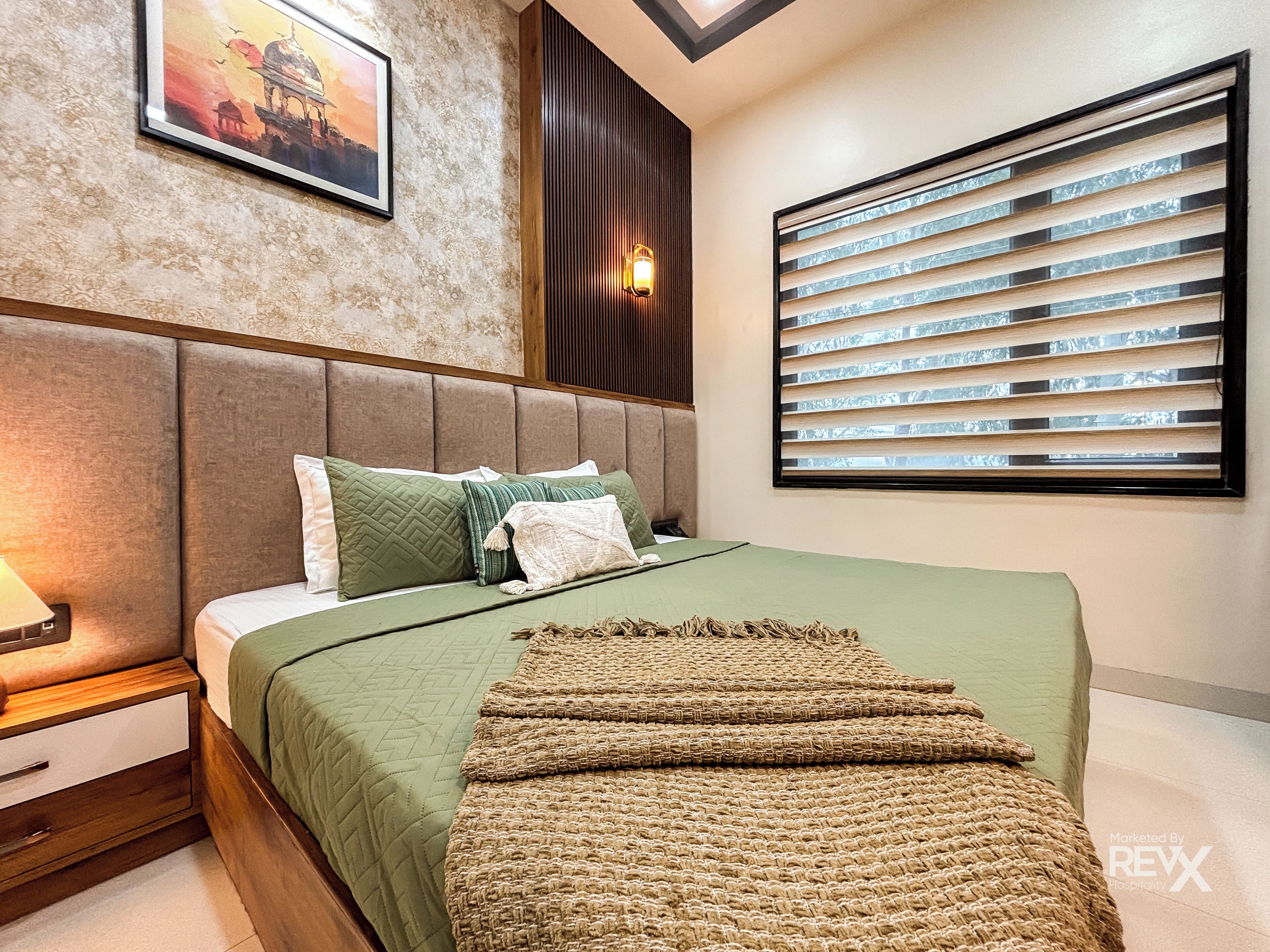 Une chambre moderne avec un grand lit, une table de chevet, une lampe fixée au mur et une fenêtre avec des stores.