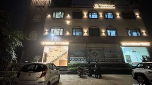 A building with illuminated signage for rooms and rooftop restaurant, a flat entrance, and a paved area with parked cars and motorcycles. - Hotel Brij Vas (Mathura)