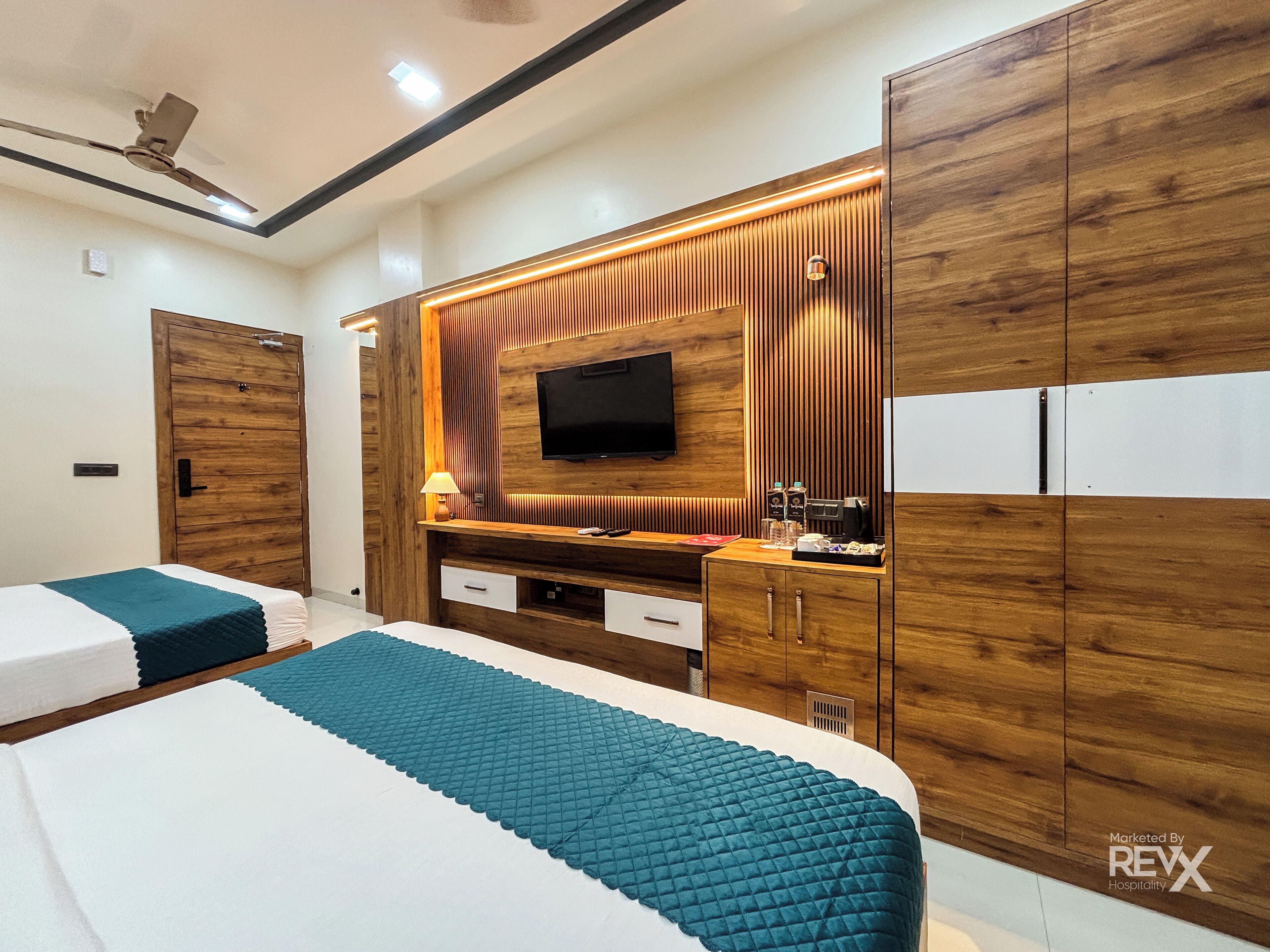 A modern hotel room with a large bed, a TV mounted on a wooden panel wall, a desk with a lamp, and a wardrobe.