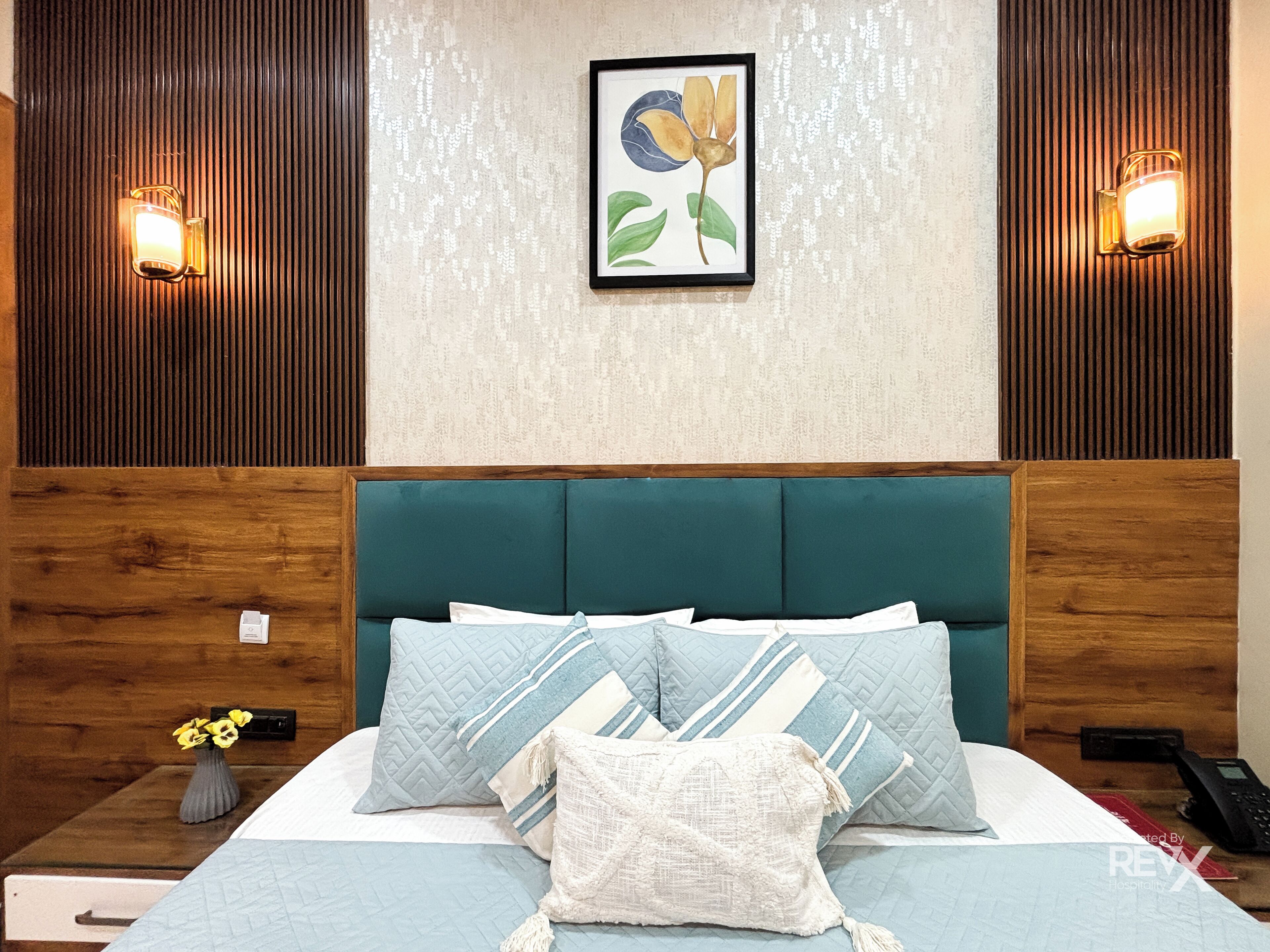 Un lit bien fait, avec une tête de lit de couleur turquoise, deux lampes fixées au mur et, au-dessus, un tableau floral encadré.