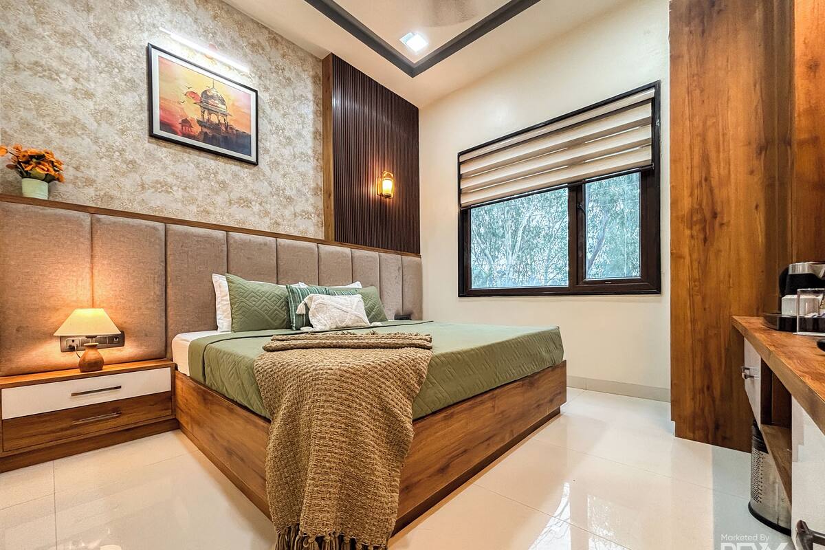 A modern bedroom with a large bed, a nightstand with a lamp, a wall-mounted picture, and a window with a view of trees.