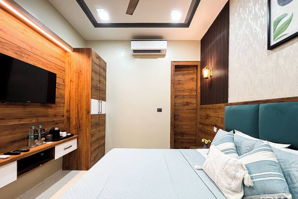 A modern bedroom with a wooden wardrobe, a flat-screen TV, a bedside table with toiletries, a bed with blue bedding, and an air conditioning unit.