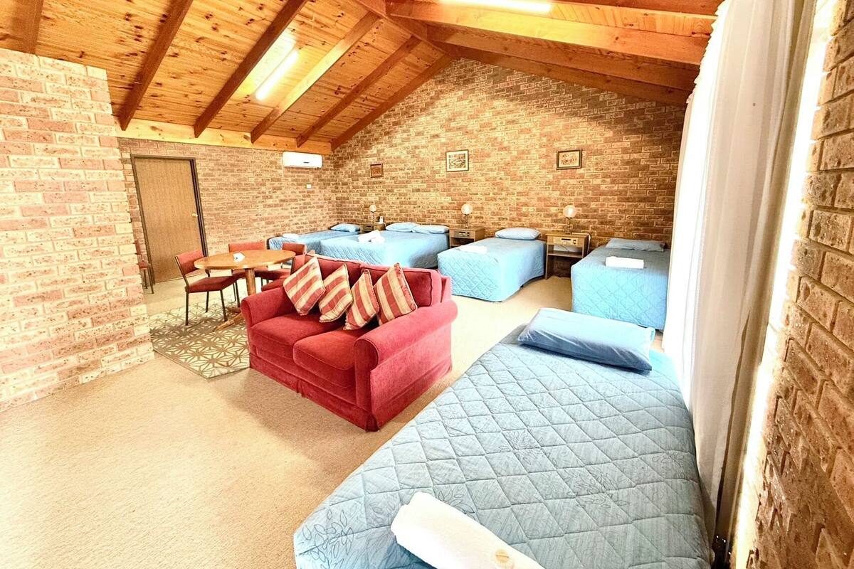 Una habitación acogedora con techo inclinado, paredes de ladrillo y vigas de madera. Cuenta con un sofá rojo, varias camas con ropa de cama azul y una pequeña mesa con sillas.