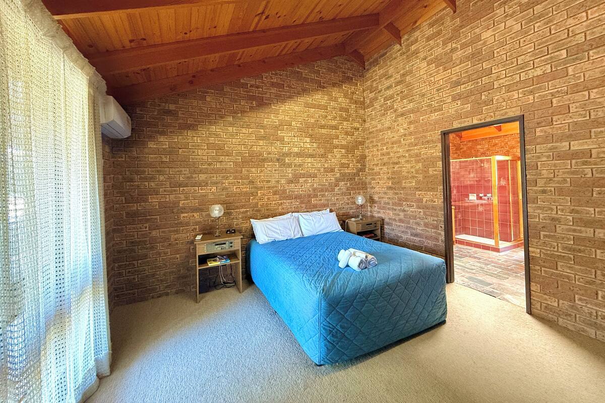 Un dormitorio con pared de ladrillo, vigas de madera en el techo, una cama con colcha azul, dos mesitas de noche y una ventana con marco rojo.