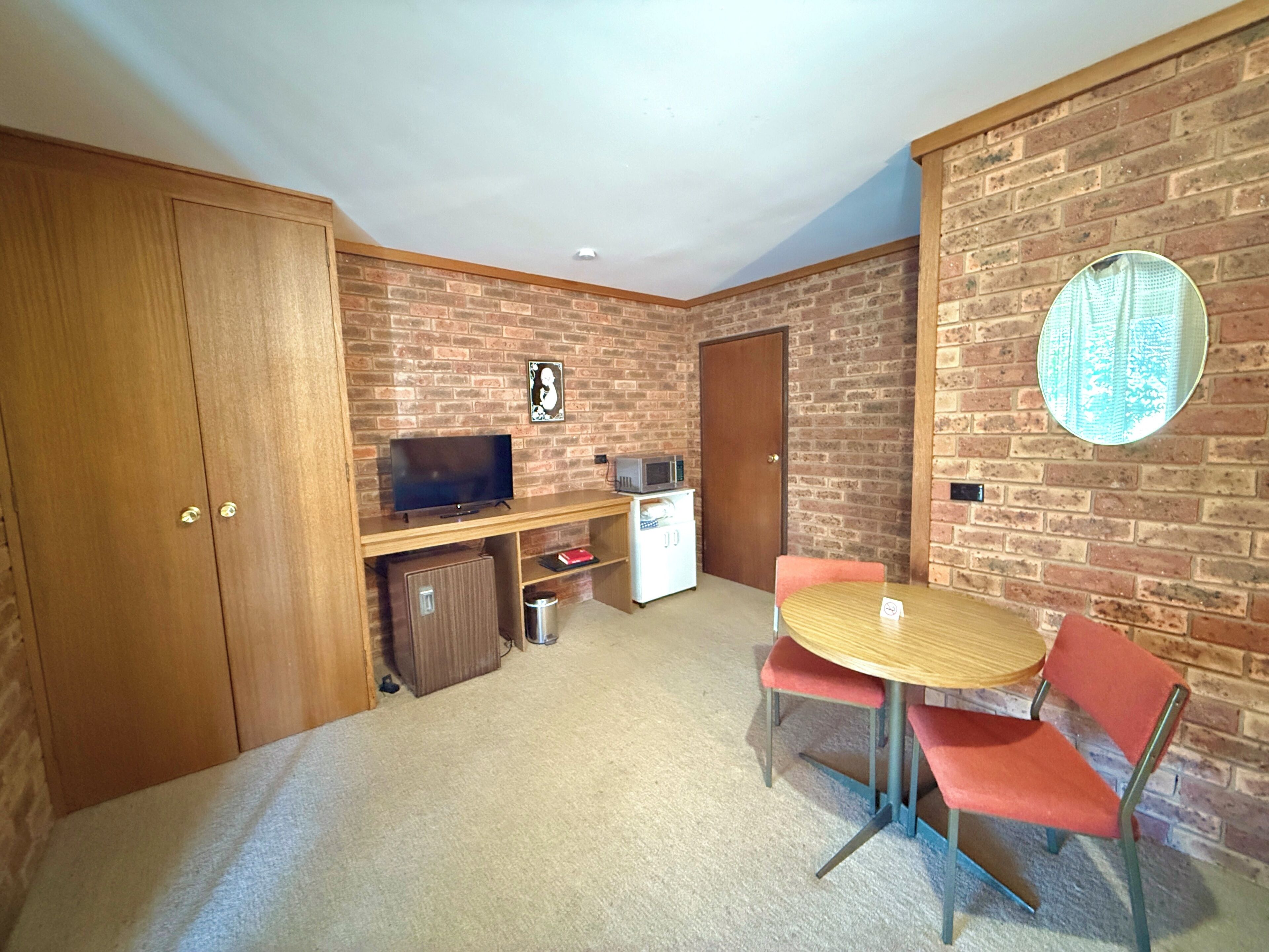 A room with a brick wall, a wooden desk with a television, a small refrigerator, and a round table with two chairs.