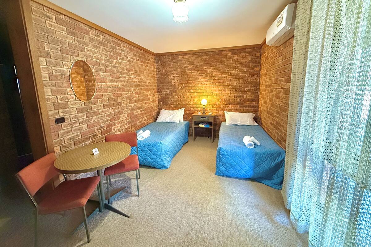Habitación pequeña con dos camas individuales, una mesita con sillas, una lámpara y una pared de ladrillo.