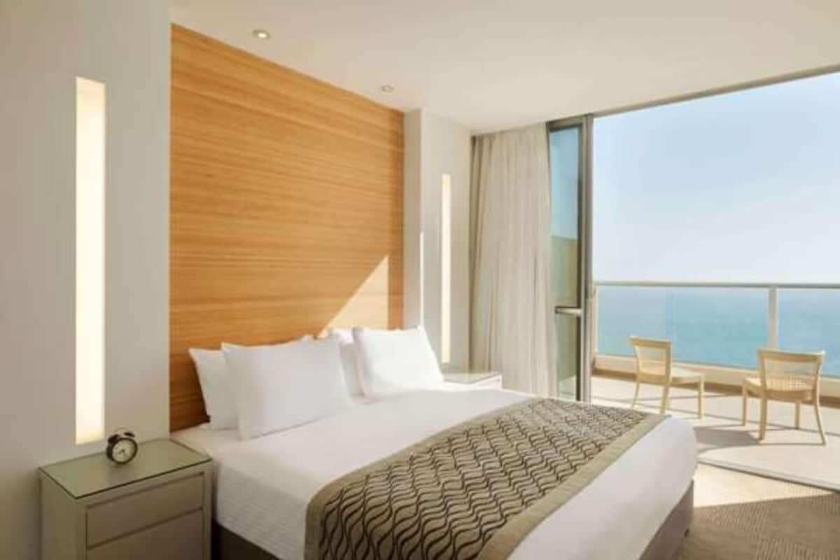 A hotel room with a large bed, a wooden headboard, a nightstand with a clock, and a balcony with chairs.