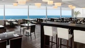 Un restaurante con vistas al océano, con piso de madera, taburetes de bar blancos e iluminación colgante.