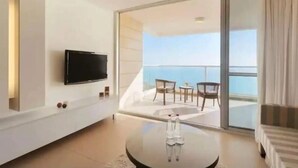 Habitación de hotel moderna con una televisión de pantalla plana montada en la pared, una mesa redonda con un par de zapatos, y un balcón con vistas al mar.
