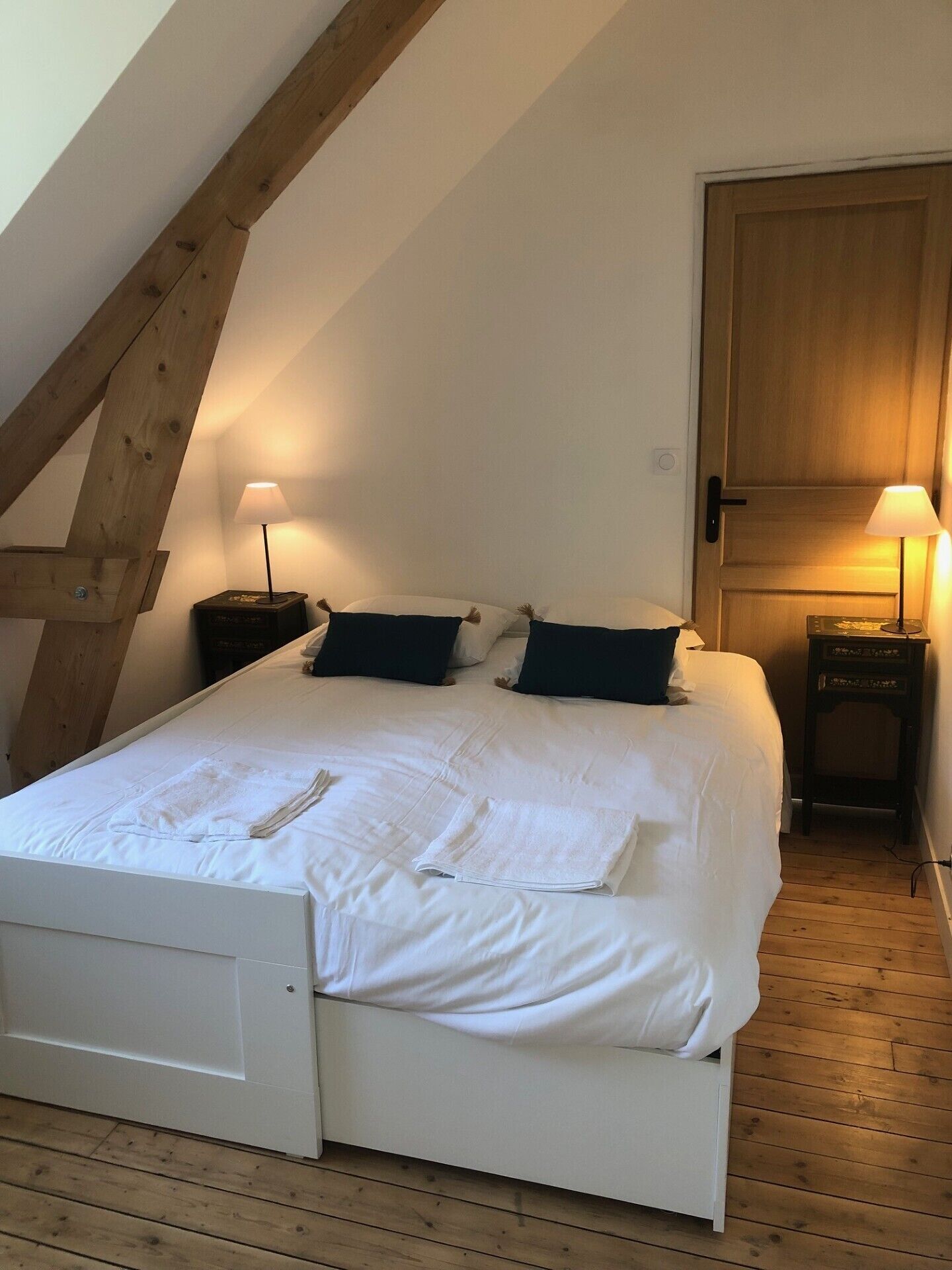 Un dormitorio con cama blanca, dos mesitas de noche con lámparas, una puerta de madera y techo inclinado.