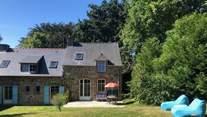A stone building with a sloped roof, multiple windows, and a door. There is a small patio with a table and chairs, and a large tree in the background. - At the heart of a market-garden farm on the Emerald Coast  (Saint-Lunaire)