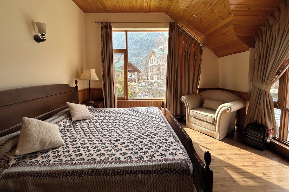 A bedroom with a wooden ceiling, a bed with patterned bedding, a chair, a lamp, and a view of a mountain and buildings.