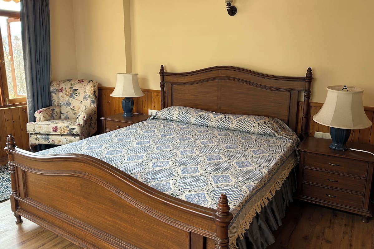 A bedroom with a wooden bed, two bedside tables with lamps, a floral armchair, and a wall-mounted light fixture.