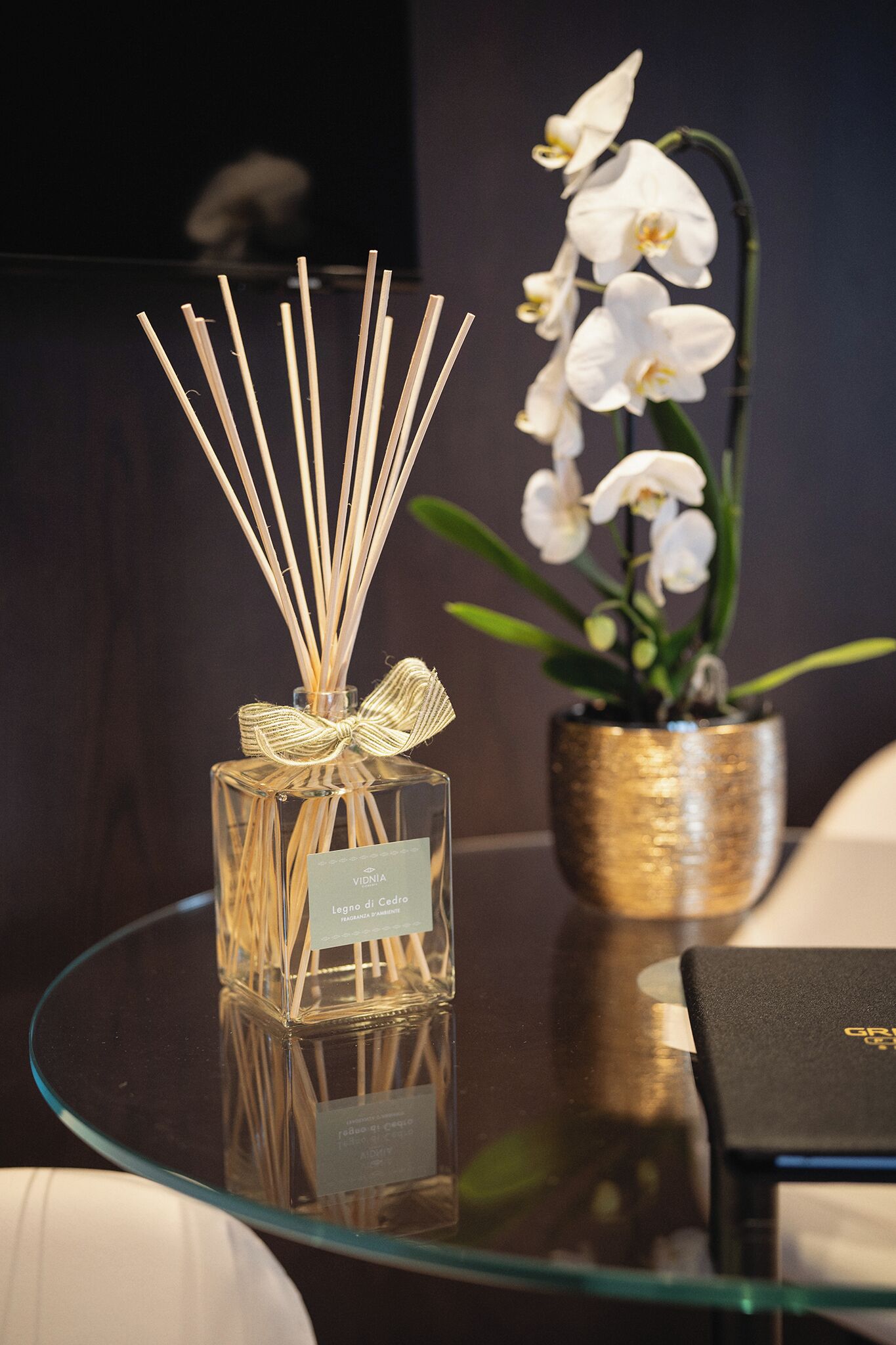 Una mesa de vidrio con un difusor de aroma de juncos y una orquídea en maceta.