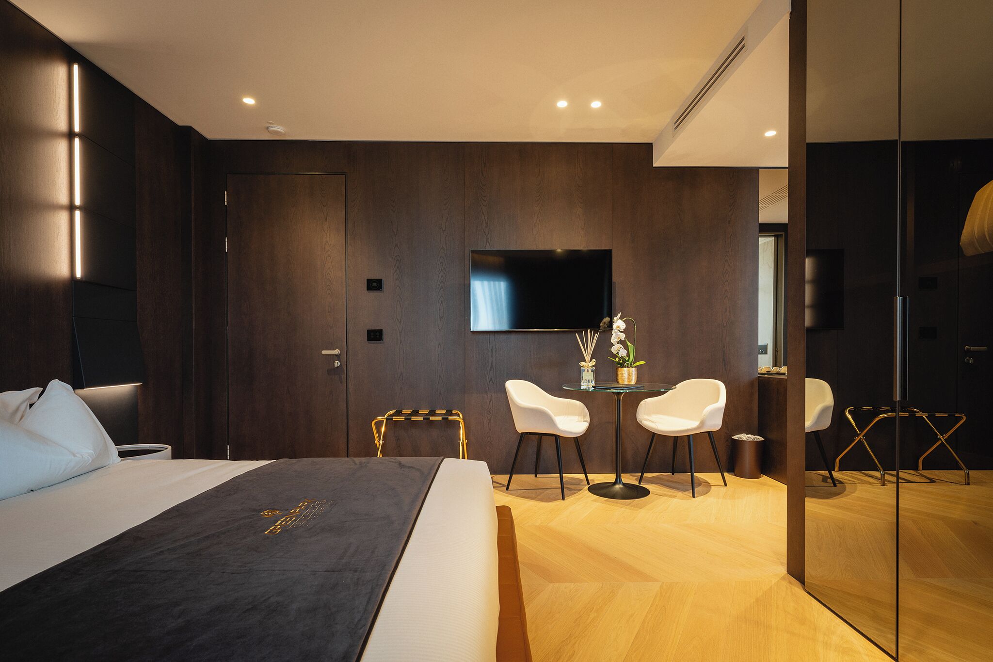 A modern hotel room with a large bed, a flat-screen TV, a small table with chairs, and a glass partition.