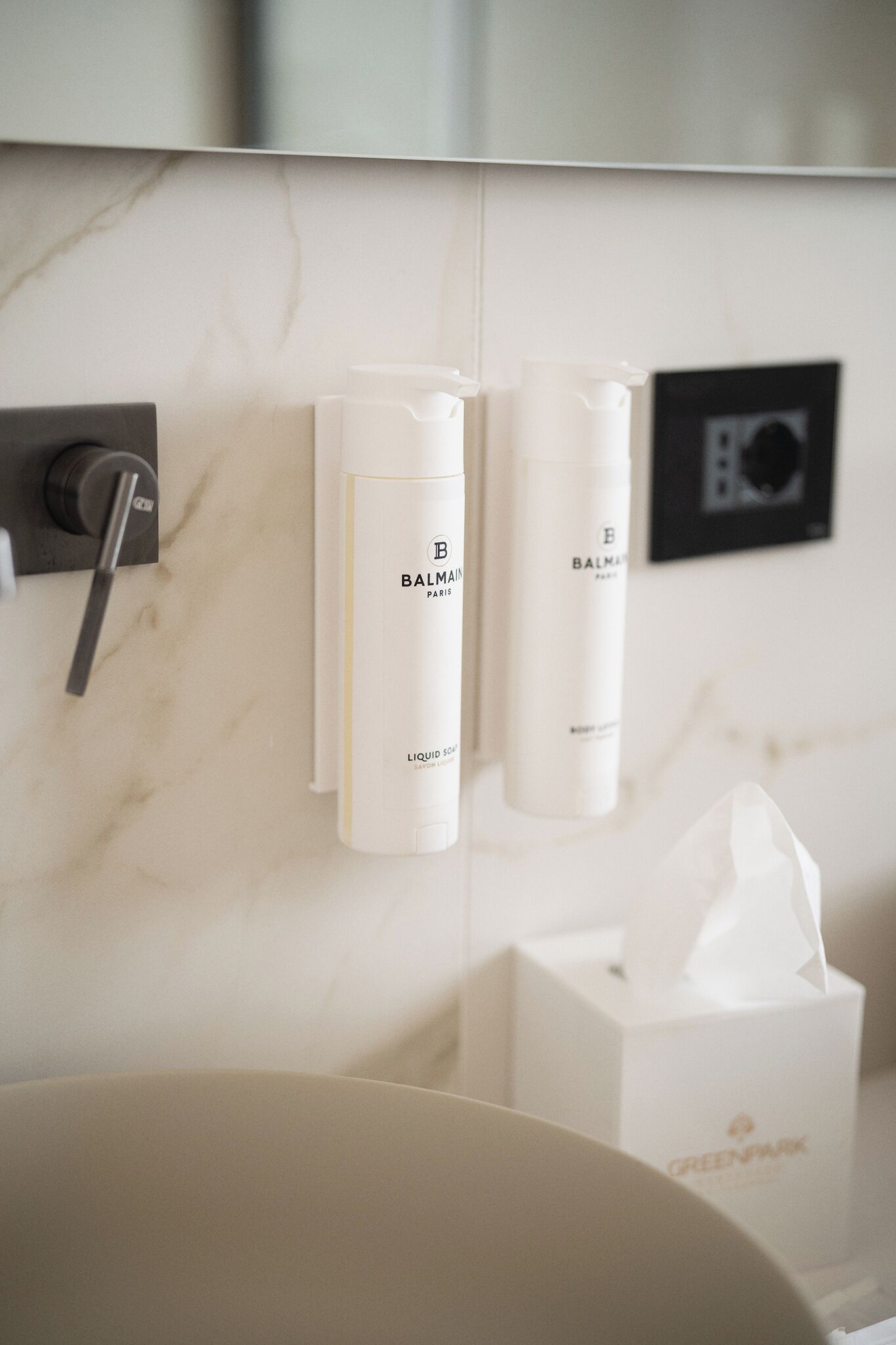 A bathroom with a marble wall, a sink, and two bottles of Balmain Paris liquid soap.