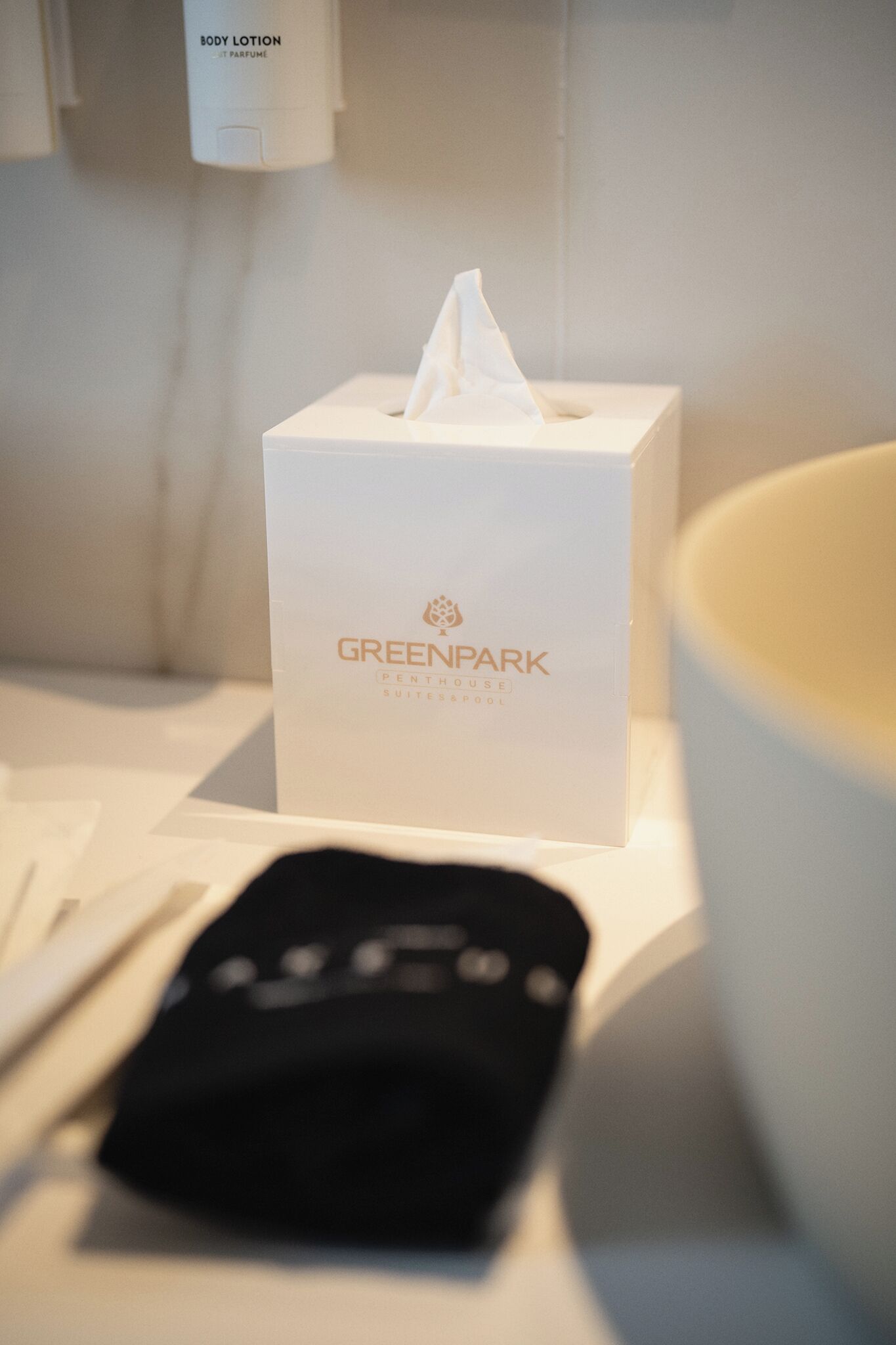 Quarto de hotel com uma caixa de lenços rotulada "GREENPARK" e um controle remoto sobre a mesa.