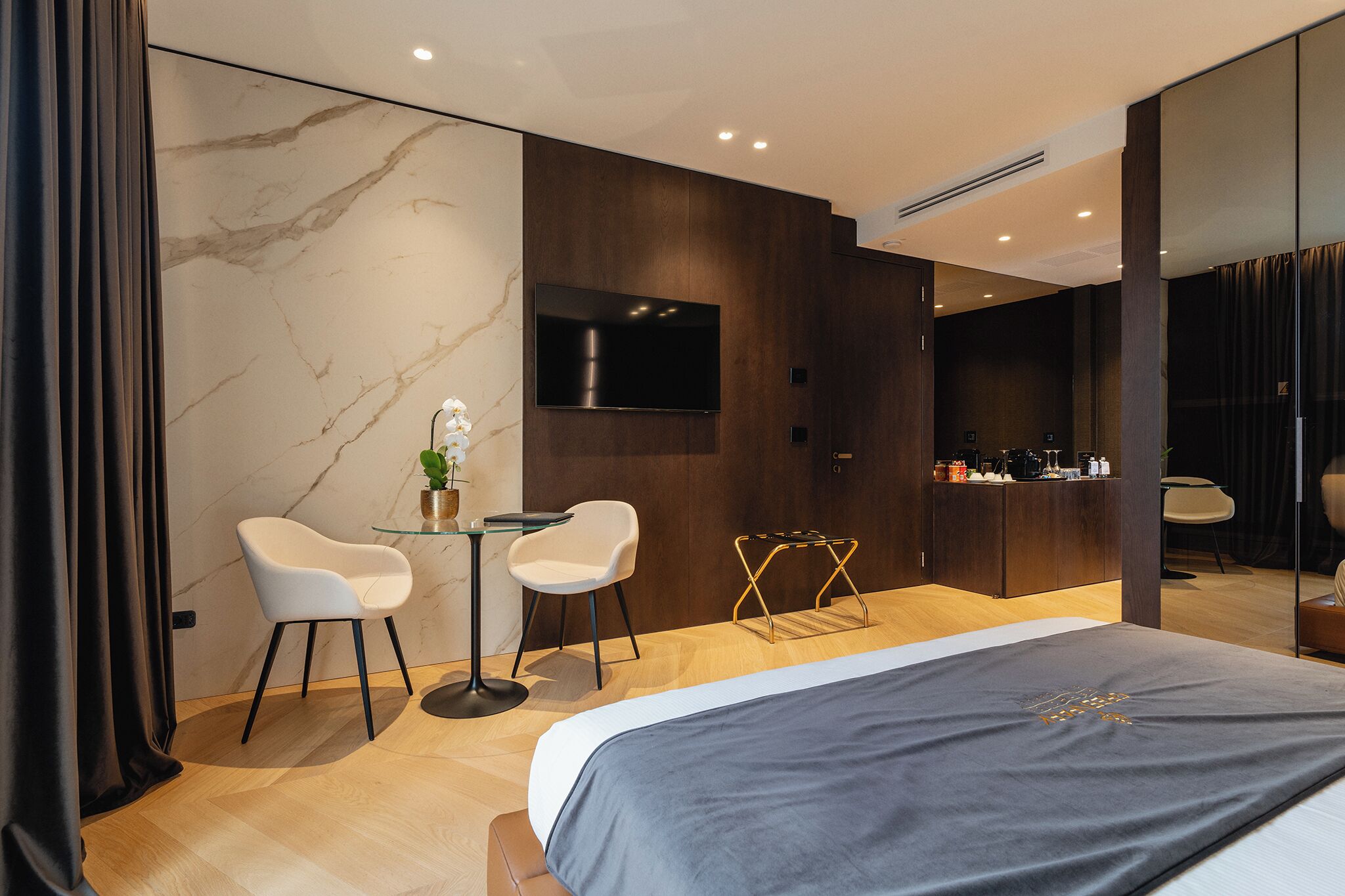 Una habitación de hotel moderna con una cama grande, dos sillas blancas, una mesita con una planta, un televisor montado en la pared y un minibár.