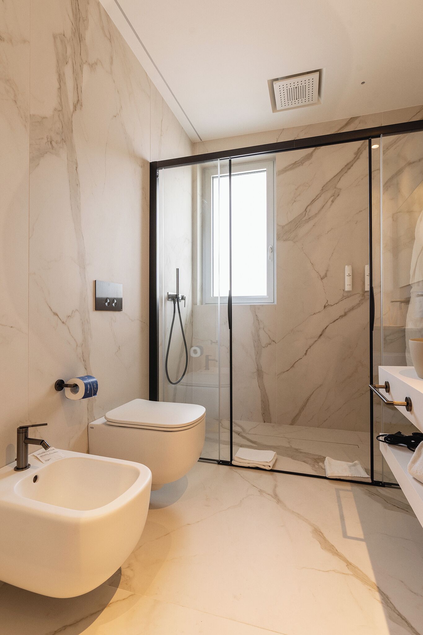 A modern bathroom with a marble floor, a freestanding bathtub, a glass-enclosed shower, and a window providing natural light.