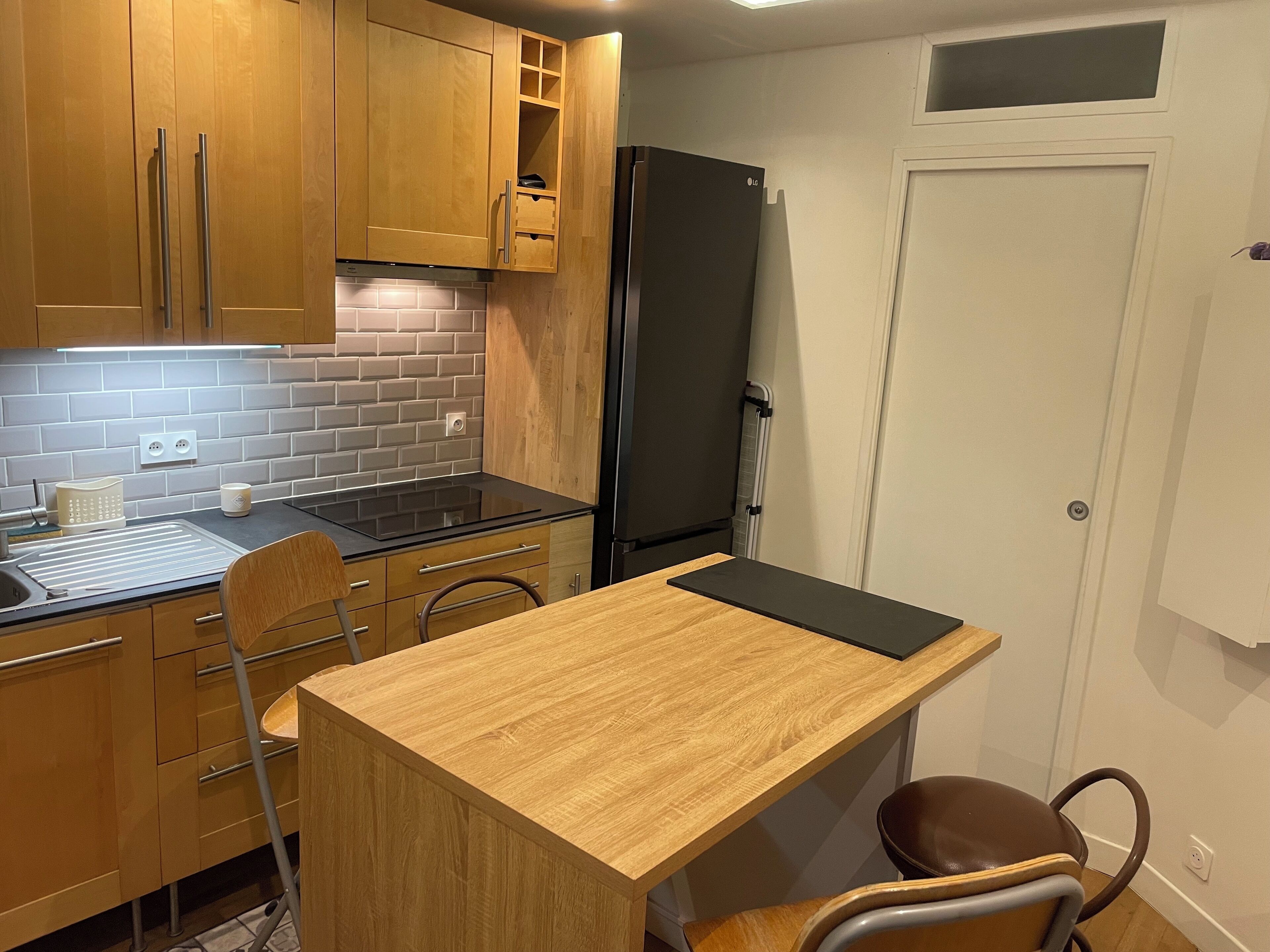 A compact kitchen with wooden cabinets, a black refrigerator, a small countertop, and a chair.