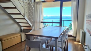 A dining area with a wooden table and white chairs, a bench, and a staircase leading to a balcony with a view of the sea.