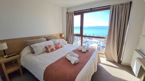 A hotel room with a large bed, a nightstand, a window with a view of the sea, and a view of the city.