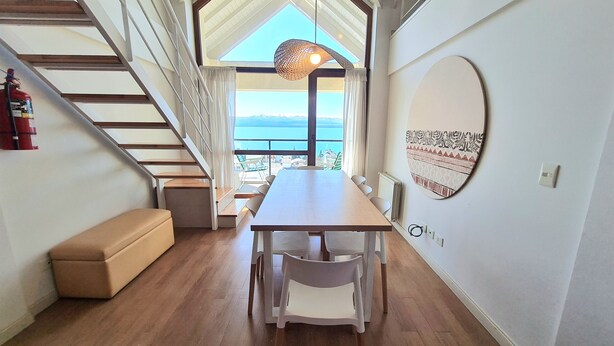 A modern interior with a wooden staircase, a dining table with chairs, a bench, and a view of the sea through large windows.