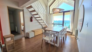 A spacious room with a wooden floor, a dining table with chairs, a staircase, and a view of the sea.