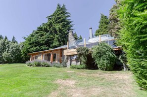 A log cabin with a stone chimney, surrounded by lush greenery and tall trees.