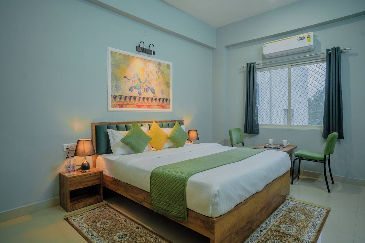 A bedroom with a wooden bed, green and yellow pillows, a green throw blanket, a nightstand with a lamp, a chair, a window with curtains, and an air conditioning unit.